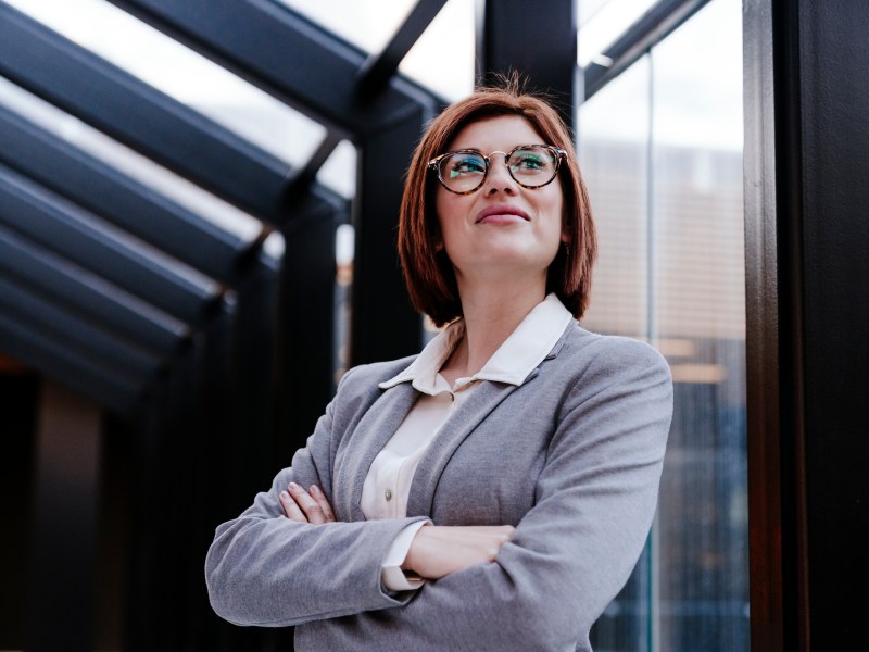 Eine Business-Frau steht mit verschrÃ¤nkten Armen vor einem Fenster.