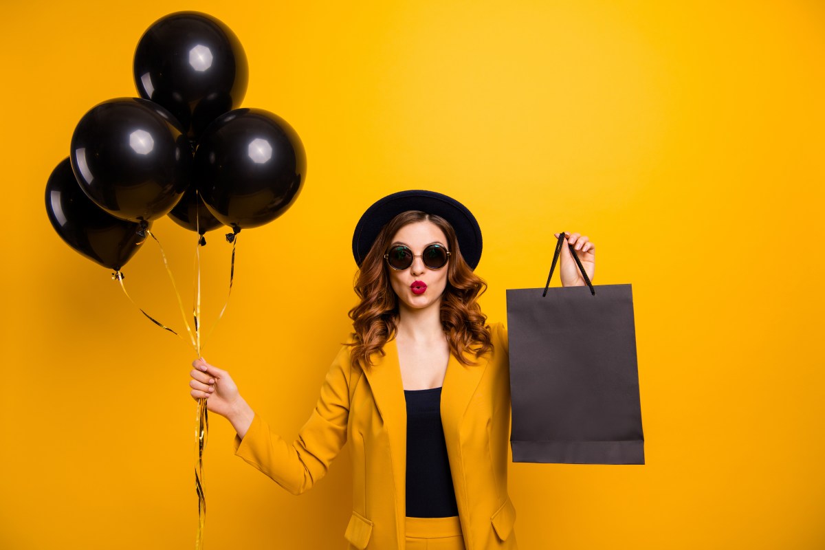 Eine Frau mit braunen Haaren und Sonnenbrille hält schwarze Luftballons und eine schwarze Tüte in der Hand.