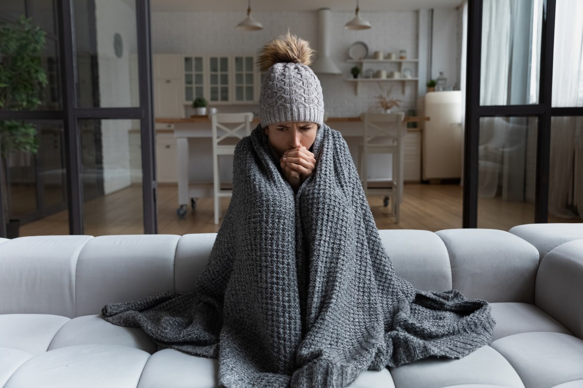 Eine Frau sitzt frierend in ihrer Wohnung. Sie trägt eine Mütze und ich in eine Decke eingewickelt.