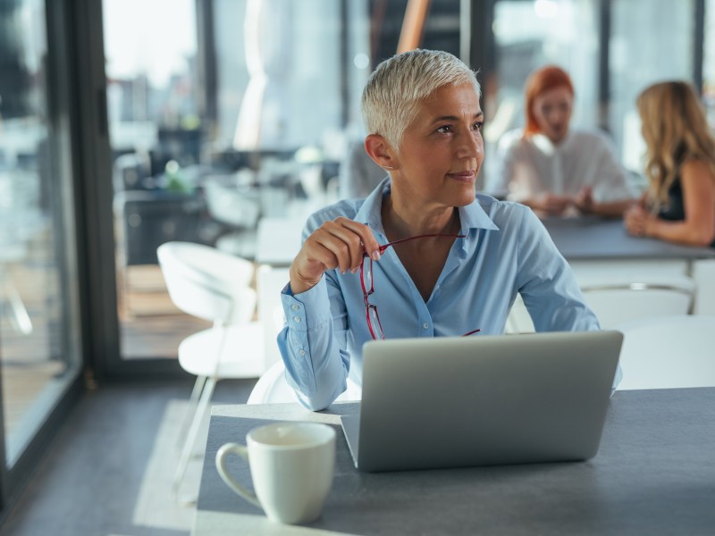 Eine ältere Dame mit grauen Haare sitzt im Büro vor ihrem Laptop.