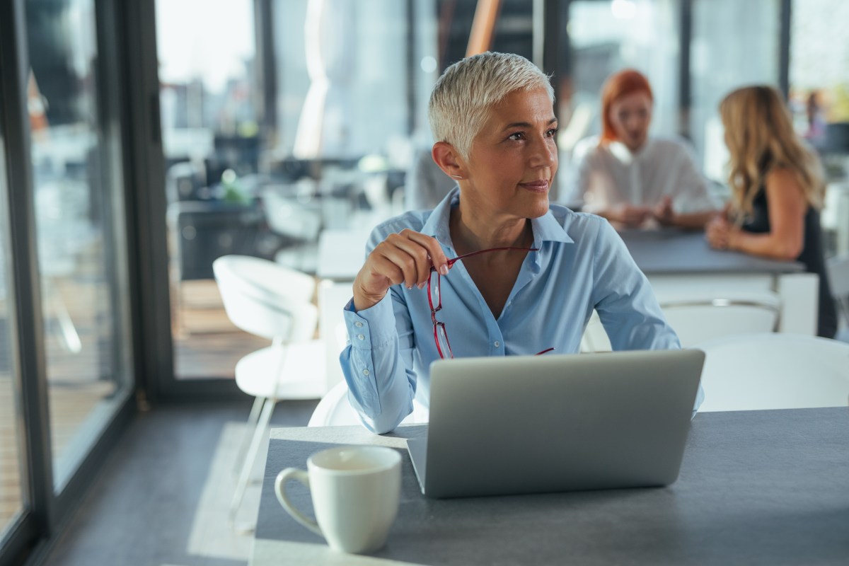 Eine ältere Dame mit grauen Haare sitzt im Büro vor ihrem Laptop.