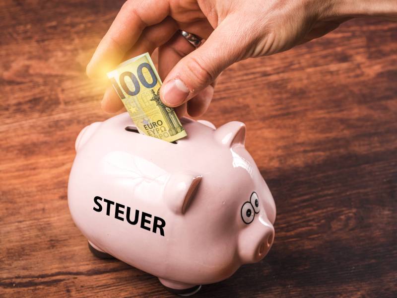 Eine Person wirft 100 Euro in ein Sparschwein mit der Aufschrift "Steuer".