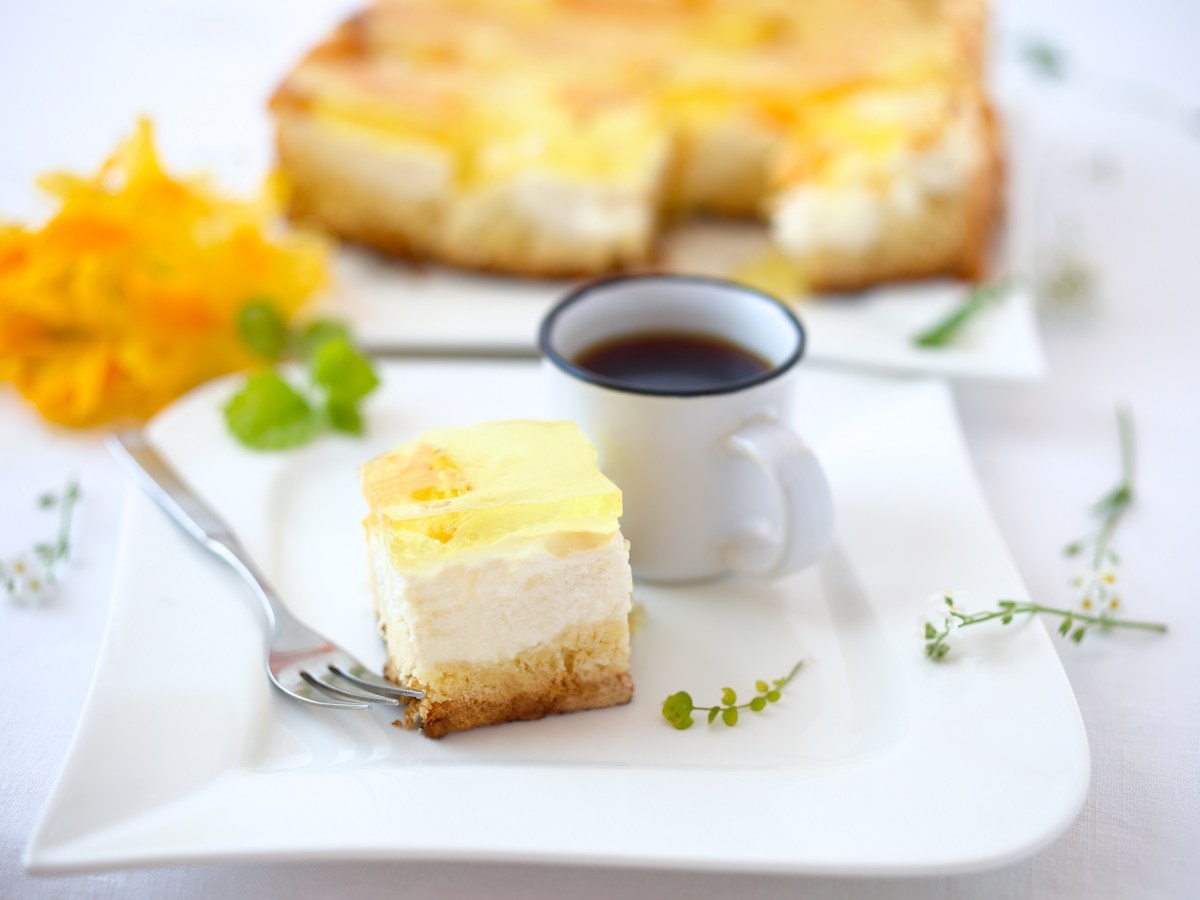 Ein StÃ¼ck Schmankuchen vom Blech mit FrÃ¼chten auf einem Teller, daneben eine Kaffeetasse, dahinter der Kuchen.