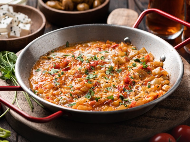 Türkisches Rührei Menemen in einer Pfanne. Dahinter stehen Schalen mit Zutaten und Flaschen mit Öl.