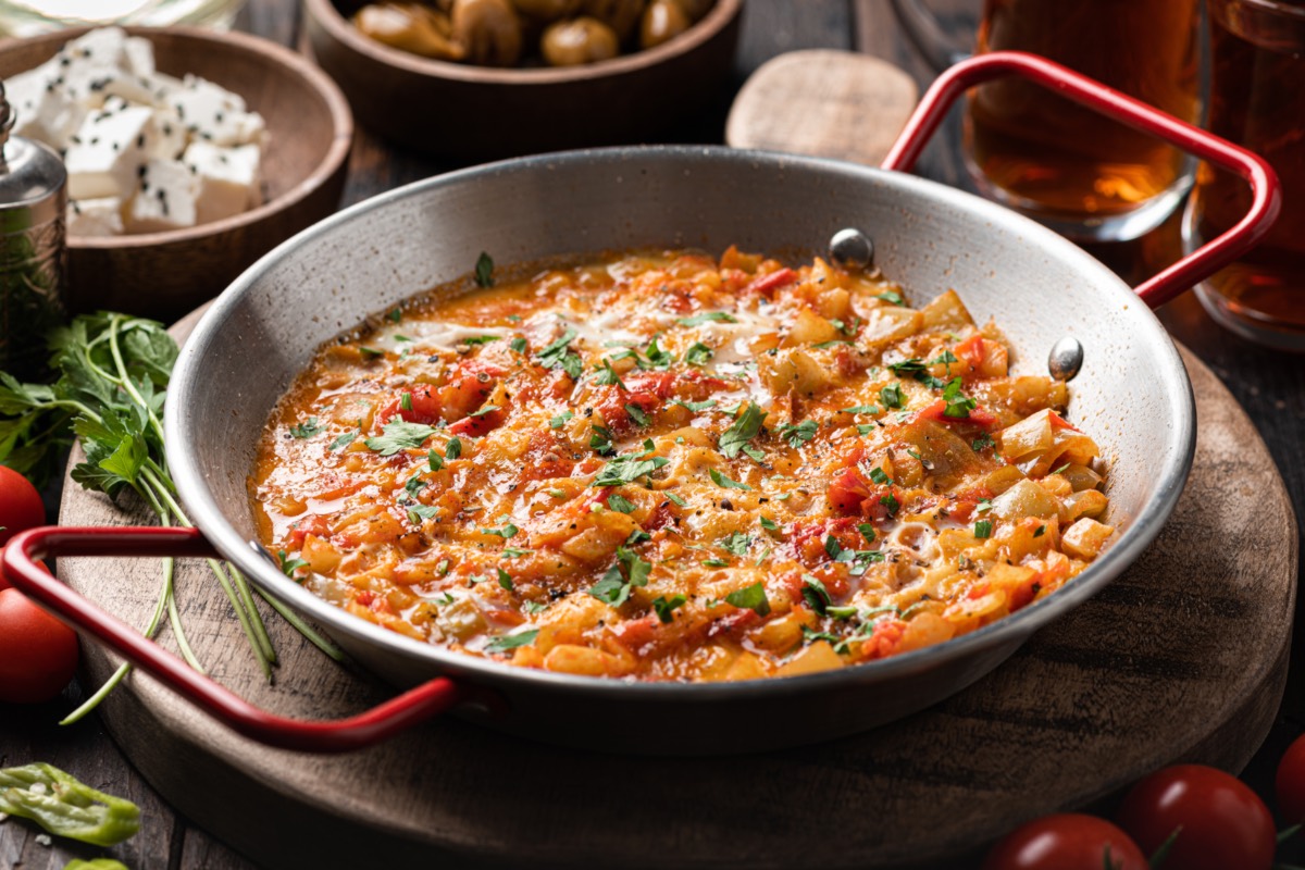 Türkisches Rührei Menemen in einer Pfanne. Dahinter stehen Schalen mit Zutaten und Flaschen mit Öl.