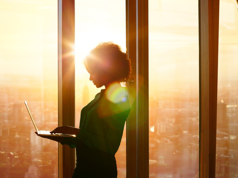 Eine Frau steht mit ihrem Laptop am Fenster. Die Sonne geht gerade unter.