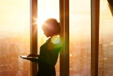 Eine Frau steht mit ihrem Laptop am Fenster. Die Sonne geht gerade unter.