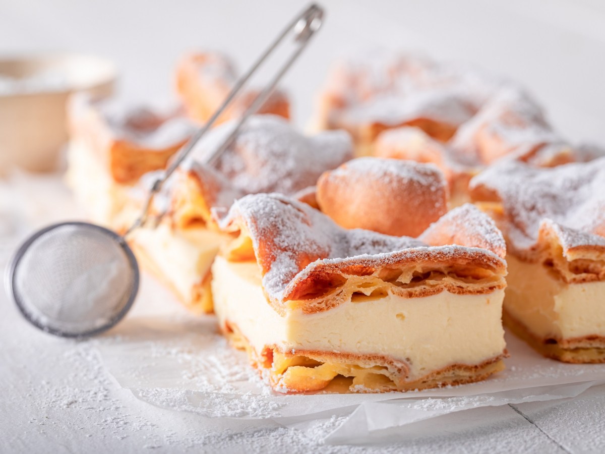 BlÃ¤tterteigkuchen mit Vanillepudding auf einer weiÃŸen Marmorplatte. Darauf liegt ein Sieb, mit dem man den Kuchen mit Puderzucker bestreuen kann.