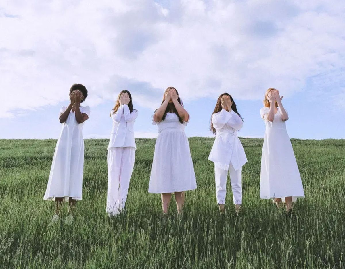 Embodiment: Frauen stehen auf einer Wiese und halten sich die Hände vors Gesicht.