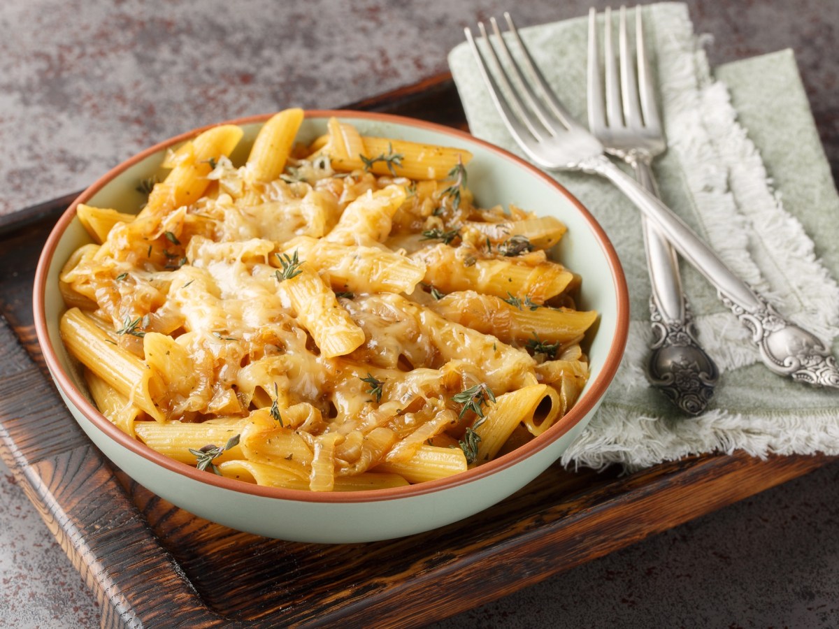 French Onion Pasta mit Zwiebeln und Penne auf einem Teller, der auf einem Brett steht daneben zwei Gabeln und Stoffservietten.