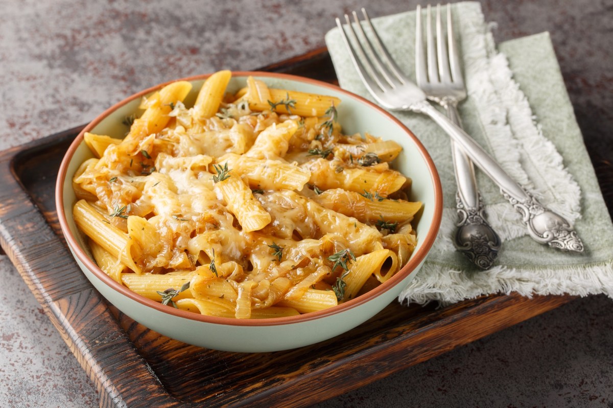 French Onion Pasta mit Zwiebeln und Penne auf einem Teller, der auf einem Brett steht daneben zwei Gabeln und Stoffservietten.