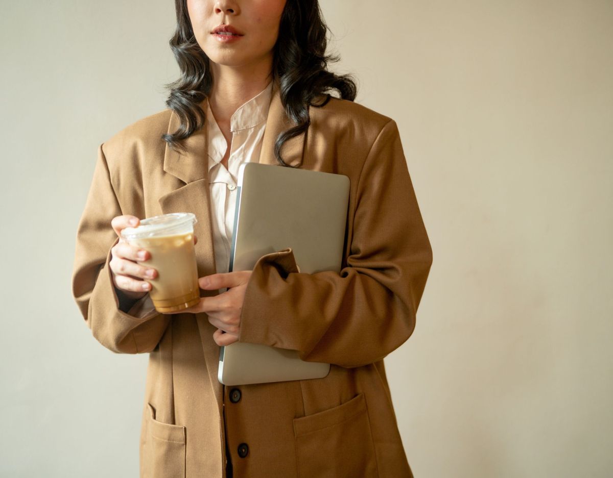 Eine Frau im beigen Mantel hält ihren Laptop und einen Eiskaffee in der Hand.