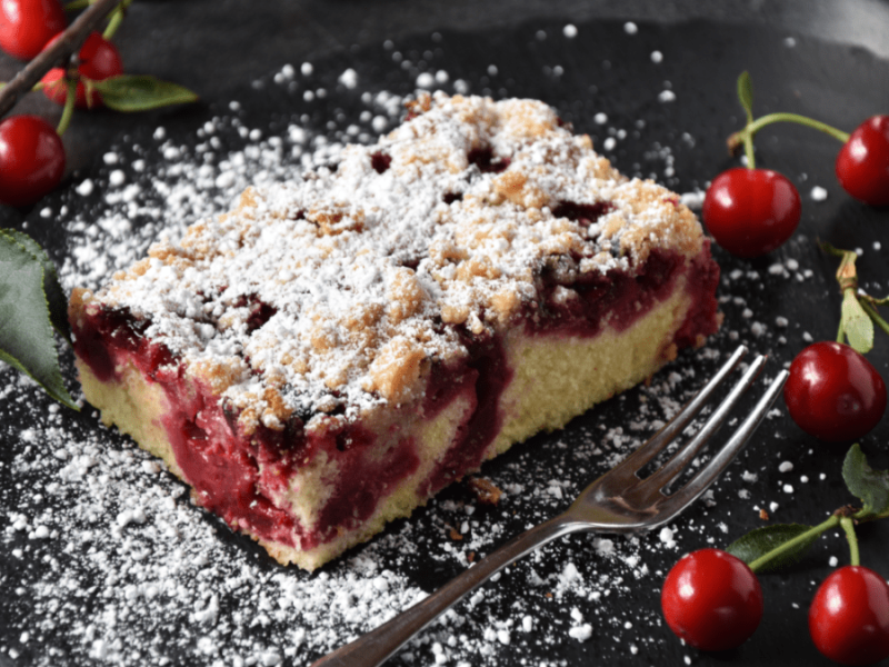 Ein Stück Kirsch-Streuselkuchen vom Blech mit Puderzucker, daneben eine Gabel und frische Kirschen