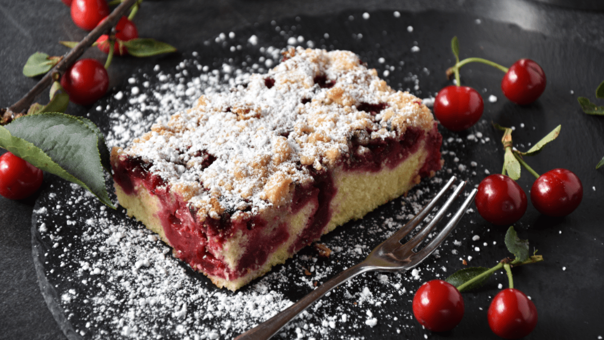 Ein Stück Kirsch-Streuselkuchen vom Blech mit Puderzucker, daneben eine Gabel und frische Kirschen