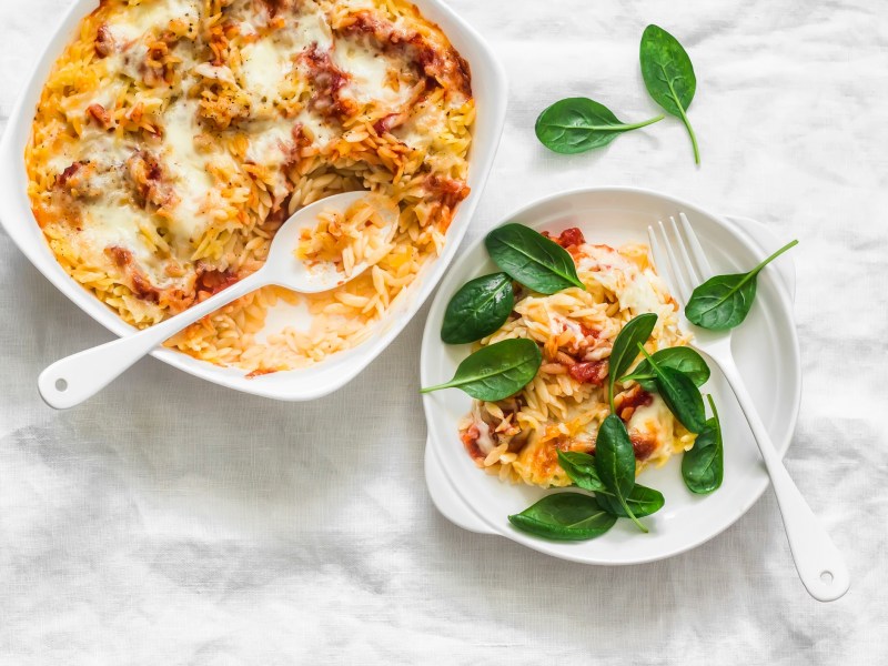 Eine Auflaufform mit Orzo-Feta-Auflauf mit Tomaten, daneben ein Teller mit etwas Auflauf und frischem Spinat.