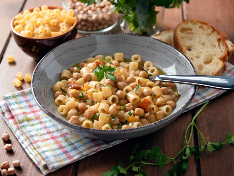 Pasta e Ceci in einer grauen SchÃ¼ssel. Daneben Schalen mit Nudeln und Kichererbsen sowie Petersilie und zwei Scheiben Brot.