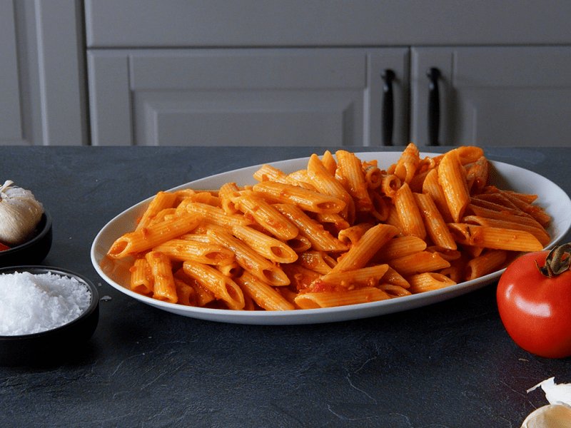 Penne Arrabiata, angerichtet auf einem Teller. Tomaten und GewÃ¼rze liegen auÃŸen herum.