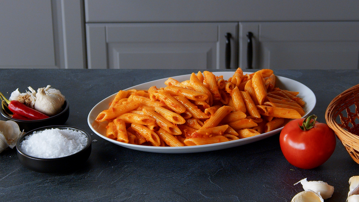 Penne Arrabiata, angerichtet auf einem Teller. Tomaten und GewÃ¼rze liegen auÃŸen herum.