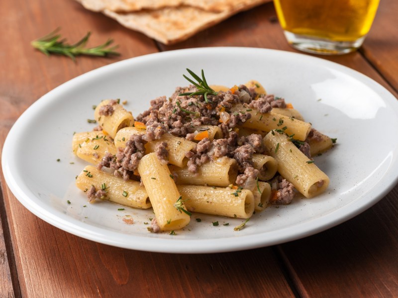 Rigatoni mit weißer Bolognese auf einem weißen Teller. Die Nudeln sind mit Kräutern dekoriert.