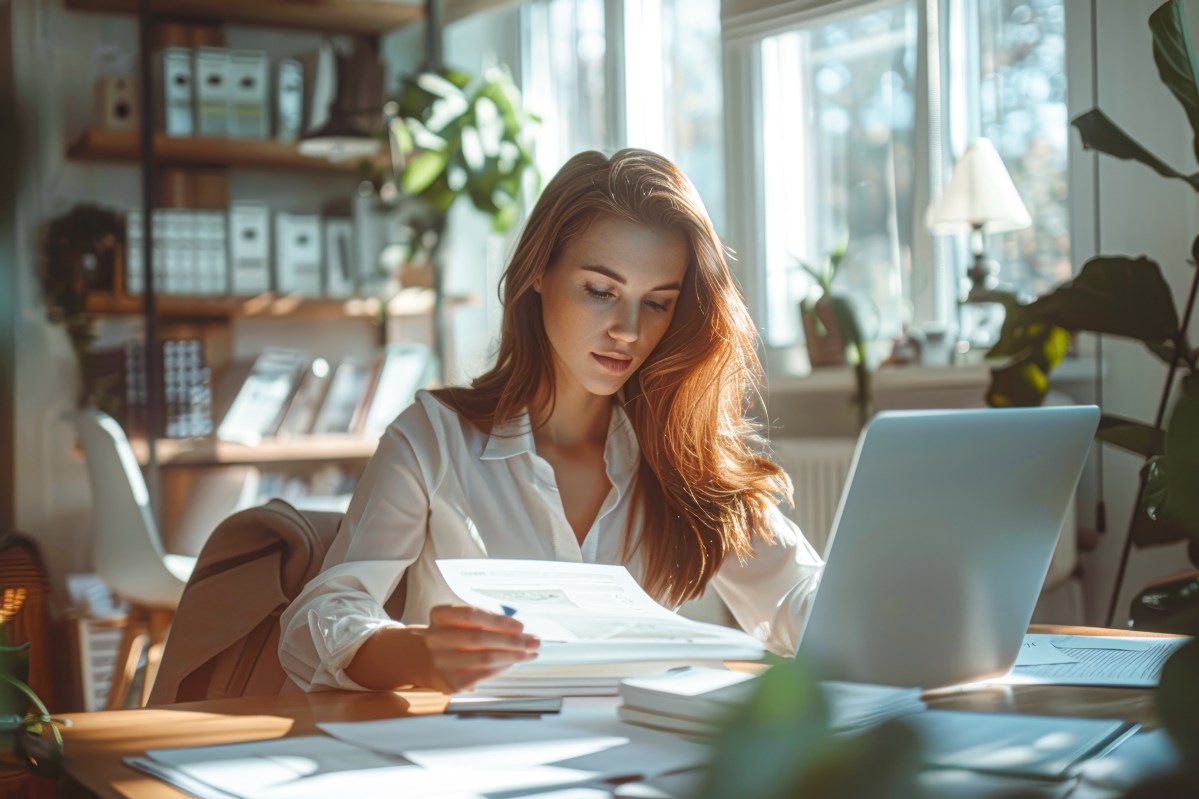 Eine Frau hält ein Dokument in der Hand. Vor ihr steht ein Laptop.