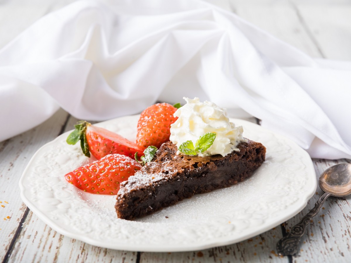 Ein StÃ¼ck schwedischer Schokoladenkuchen Kladdkaka auf einem Teller, garniert mit Erdbeeren und Sahne, daneben ein LÃ¶ffel.