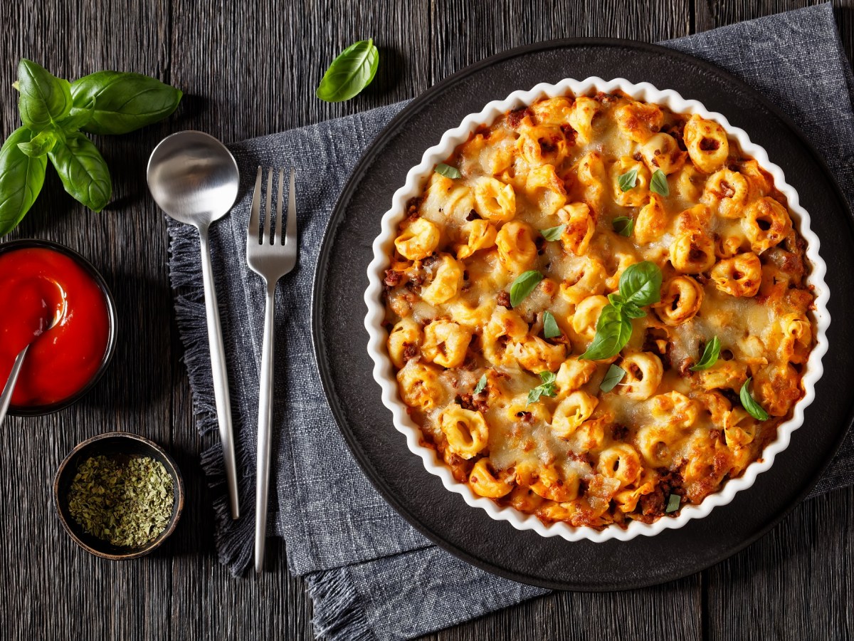 Tortellini-Auflauf in einer geriffelten Auflaufform auf einem schwarzen Tablett. Darunter ein graues Platzdeckchen, LÃ¶ffel und Gabel und TomatensoÃŸe. Draufsicht.