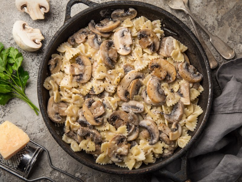 Eine gusseiserne Pfanne mit Walnuss-Pilz-Pasta in der Draufsicht, daneben ein Stück Parmesan und ein paar Champignons.