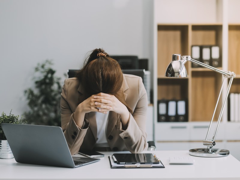 Eine junge Frau sitzt im Büro und ist erschöpft. Sie legt den Kopf auf ihre Hände.