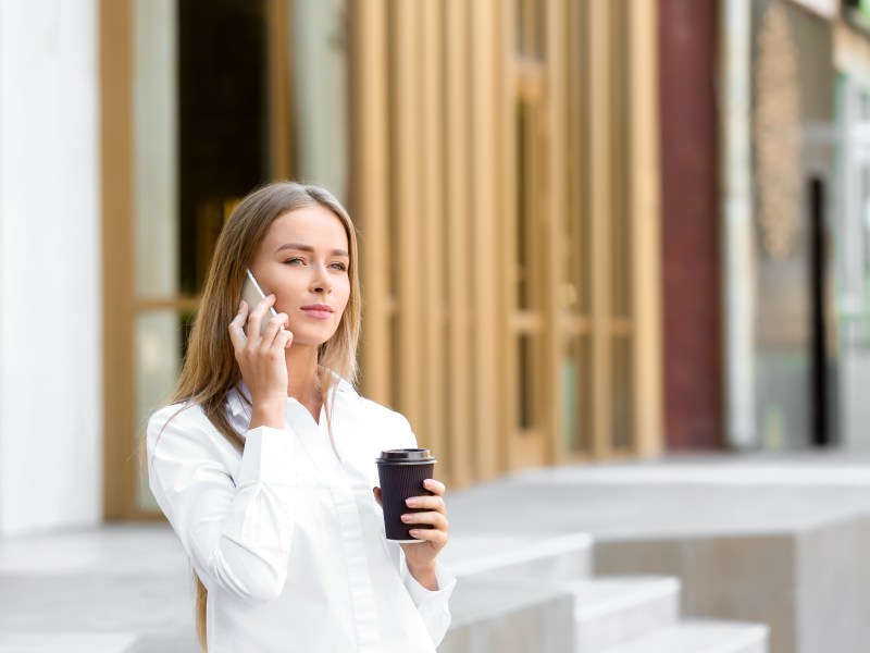 Eine Frau im weißen Hemd telefoniert und hält einen Kaffeebecher in der Hand.