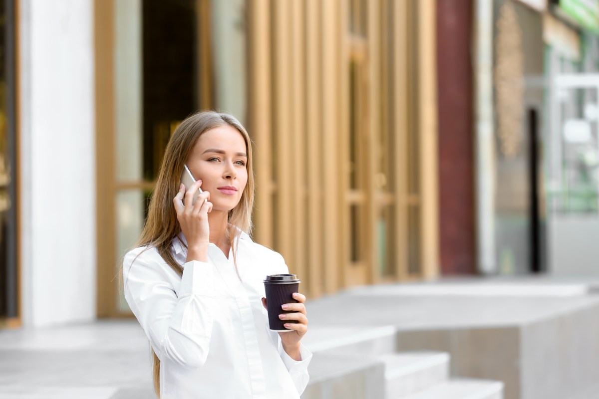 Eine Frau im weißen Hemd telefoniert und hält einen Kaffeebecher in der Hand.