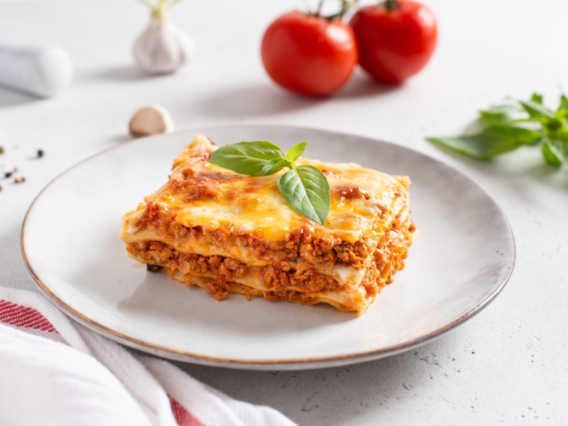 Ein Stück Lasagne mit Schmand auf einem Teller, garniert mit Basilikum, dahinter 2 Tomaten.