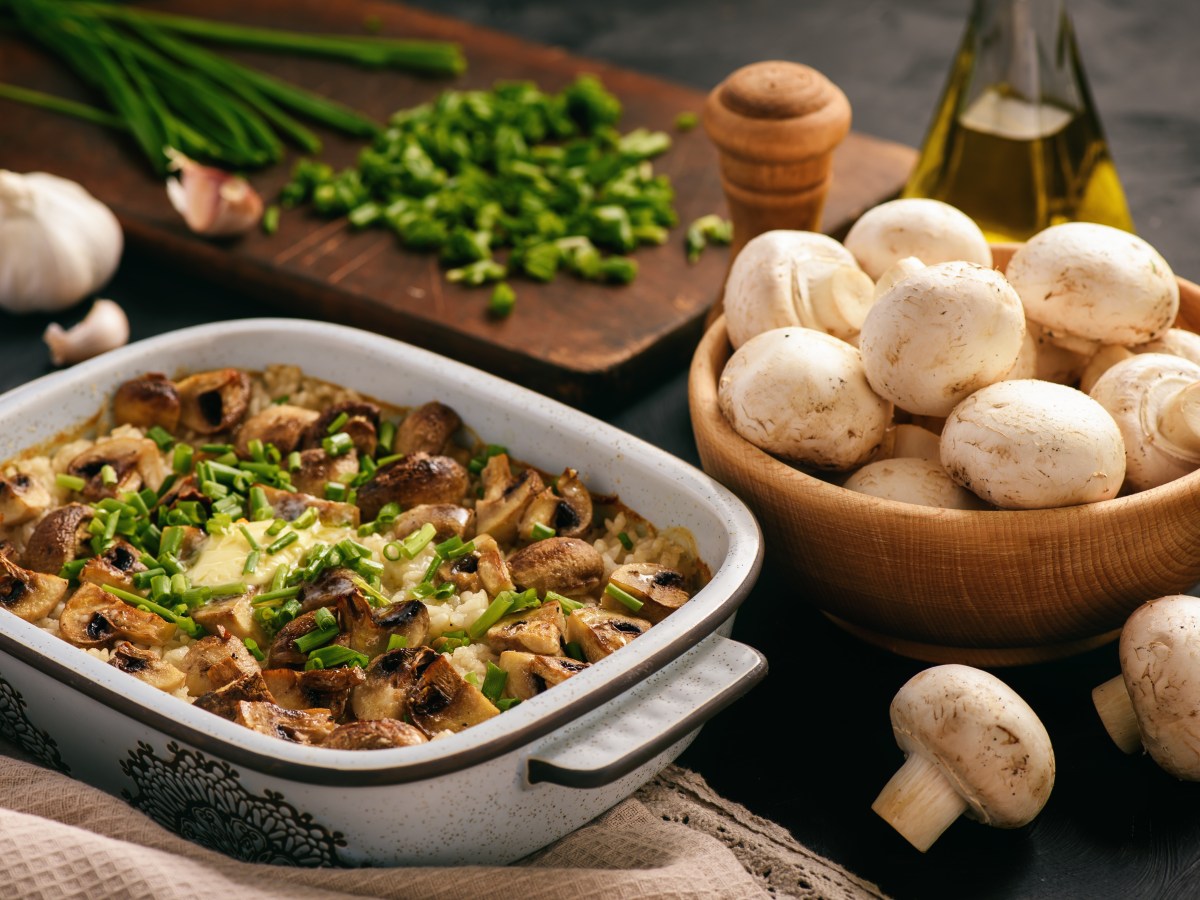 Reisauflauf mit Pilzen in Auflaufform, daneben eine Schale mit Champignons und Kräuter.