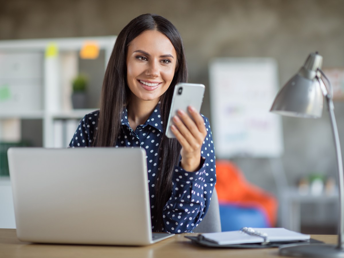 Eine Frau sitzt an ihrem Arbeitsplatz und schaut auf ihr Handy.