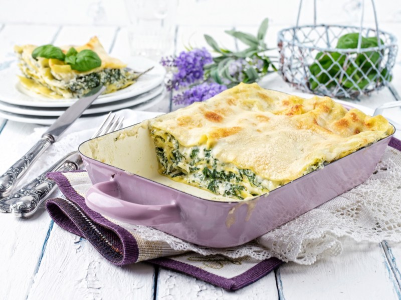 Spinat-Ricotta-Lasagne in einer Auflaufform, ein Stück bereits herausgenommen und auf einem Teller im Hintergrund.