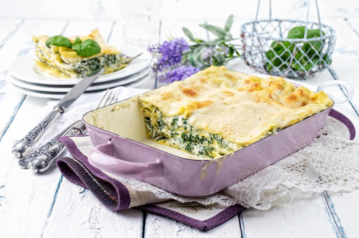 Spinat-Ricotta-Lasagne in einer Auflaufform, ein Stück bereits herausgenommen und auf einem Teller im Hintergrund.