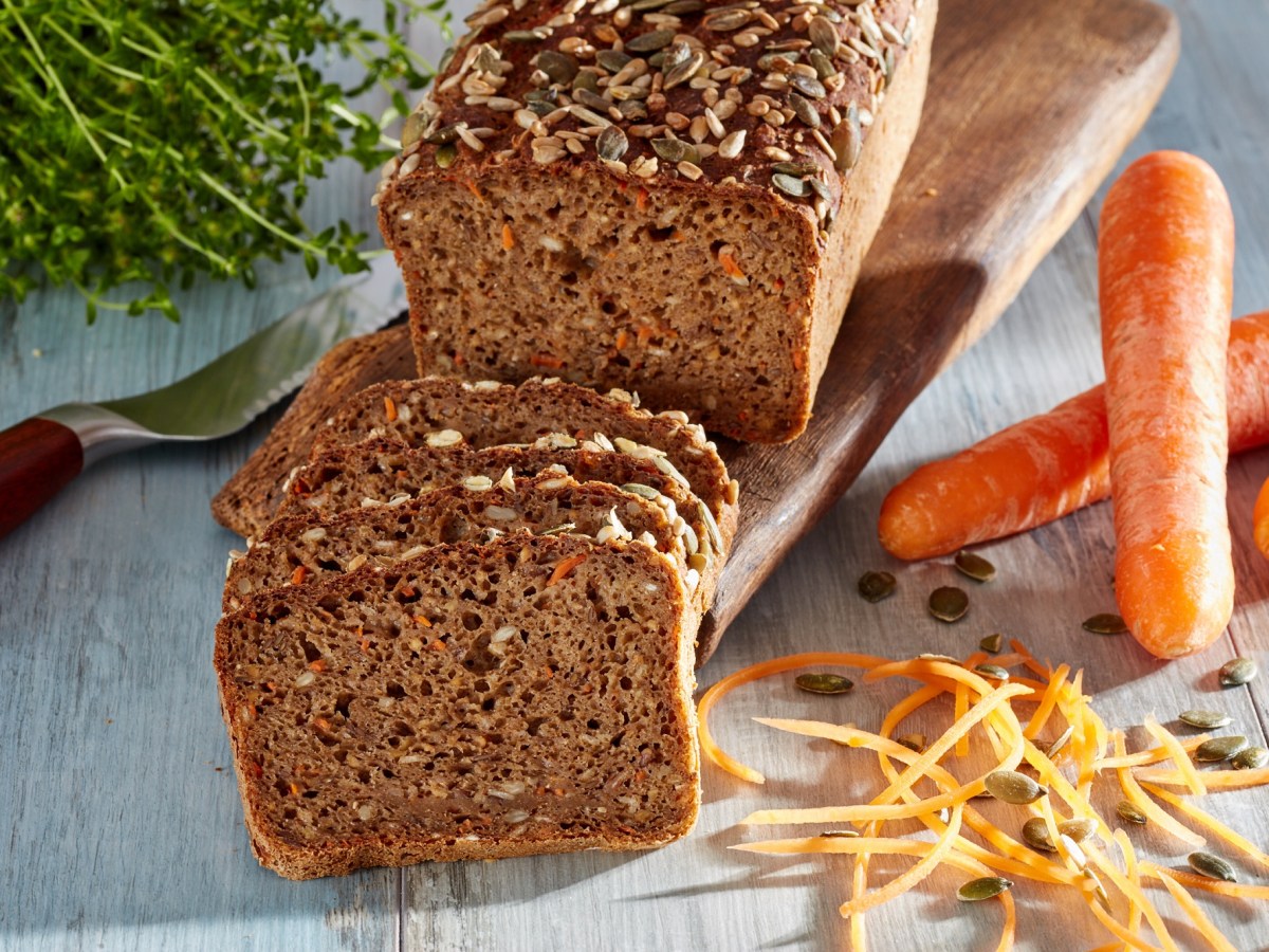 Vollkornbrot mit Möhren, angeschnitten, auf einem Holzbrett. Daneben liegen Möhren.