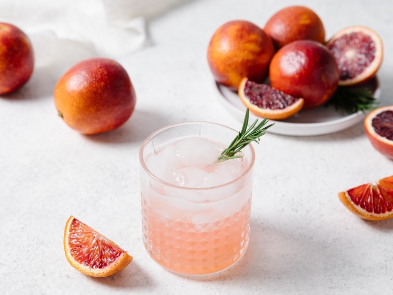 Ein Glas Aperol Paloma mit EiswÃ¼rfeln und Grapefruits daneben.