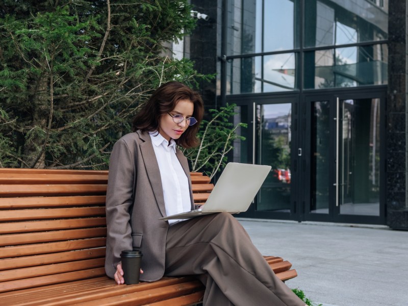 Junge Frau im Anzug benutzt Laptop auf einer Bank und schaut konzentriert.