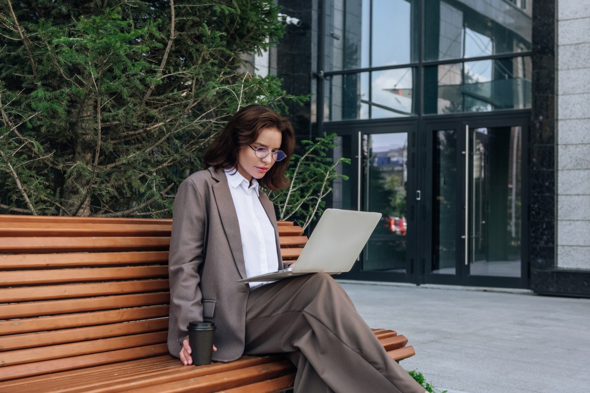 Junge Frau im Anzug benutzt Laptop auf einer Bank und schaut konzentriert.