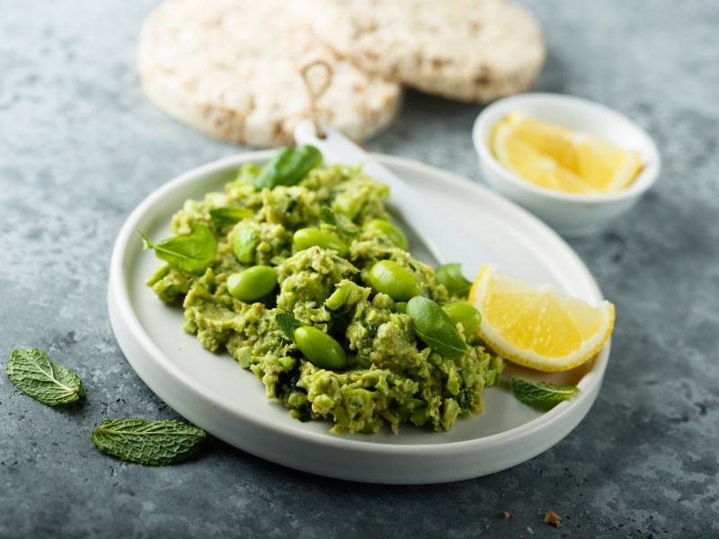 Edamame-Guacamole, mit etwas Minze garniert, auf einem weiÃŸen Teller. Serviert mit Pita-Brot.