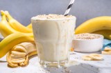 Ein Glas FrÃ¼hstÃ¼cksshake mit Bananen und Haferflocken. Im Hintergrund sind Bananen uds eine Schale mit Haferflocke zu sehen.