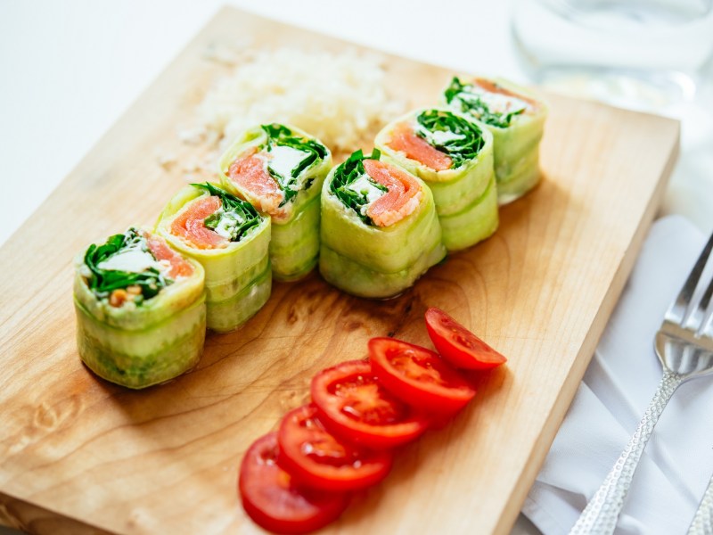 6 Gurken-RÃ¶llchen mit Lachs und Feta auf einem Brett, daneben in Scheiben geschnittene Tomaten.