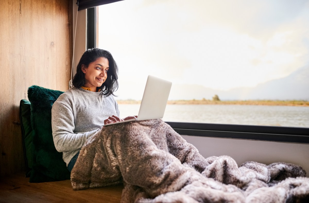 Frau sitzt mit Decke am groÃŸen Fenster und benutzt Laptop zu Hause.