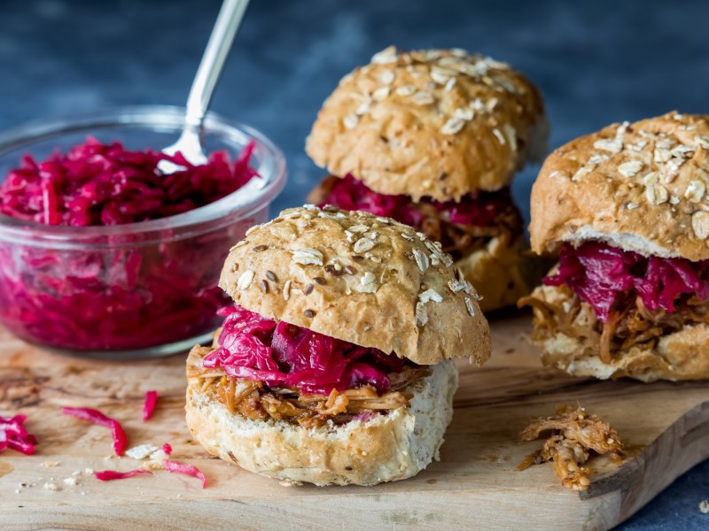 3 kleine Rotkohl-GÃ¤nse-Burger auf einem Brett, auf dem auch eine GlasschÃ¼ssel mit Rotkohl steht.