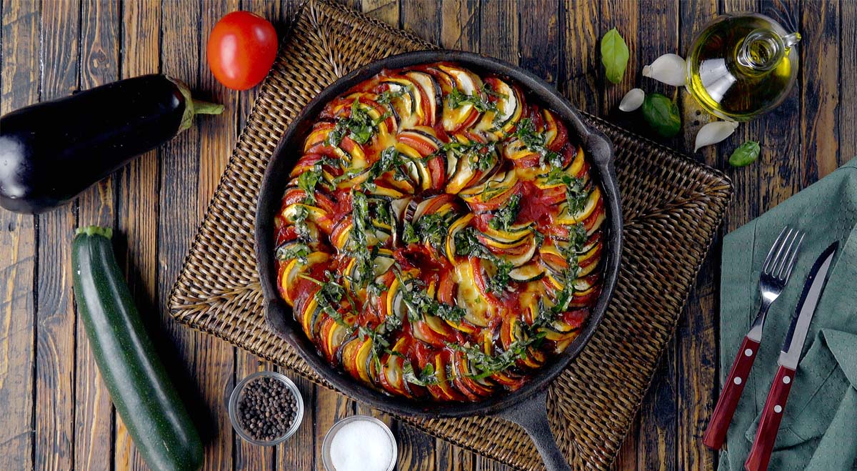 Ratatouille in der Pfanne, von oben fotografiert. Daneben liegen Besteck und frische Zutaten.