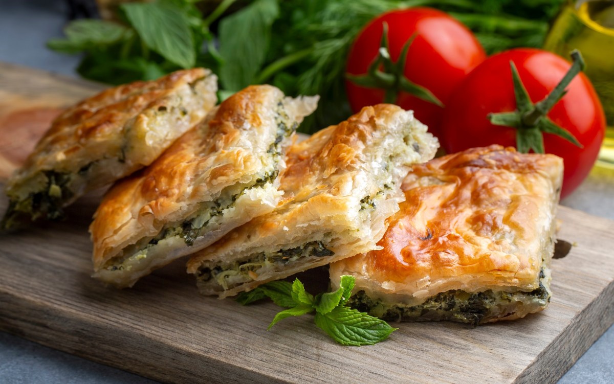 4 StÃ¼cke Spinat-BÃ¶rek mit Feta auf einem Brett, dahinter Tomaten und frischer Spinat.