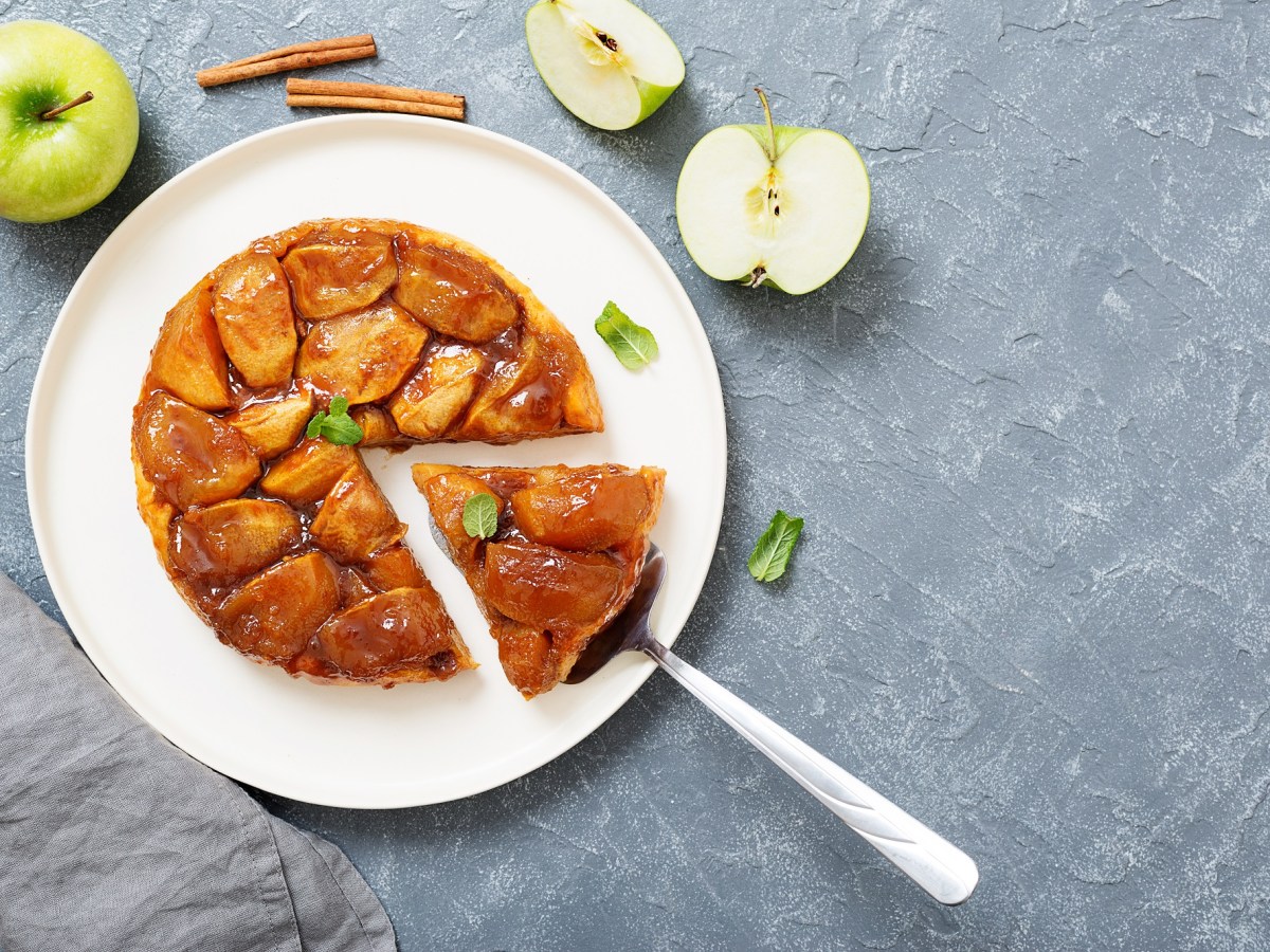 Tarte Tatin auf einem weiÃŸen Teller. Es wurde bereits ein StÃ¼ck abgeschnitten. Daneben liegen angeschnittene grÃ¼ne Ã„pfel und ein paar Zimtstangen.