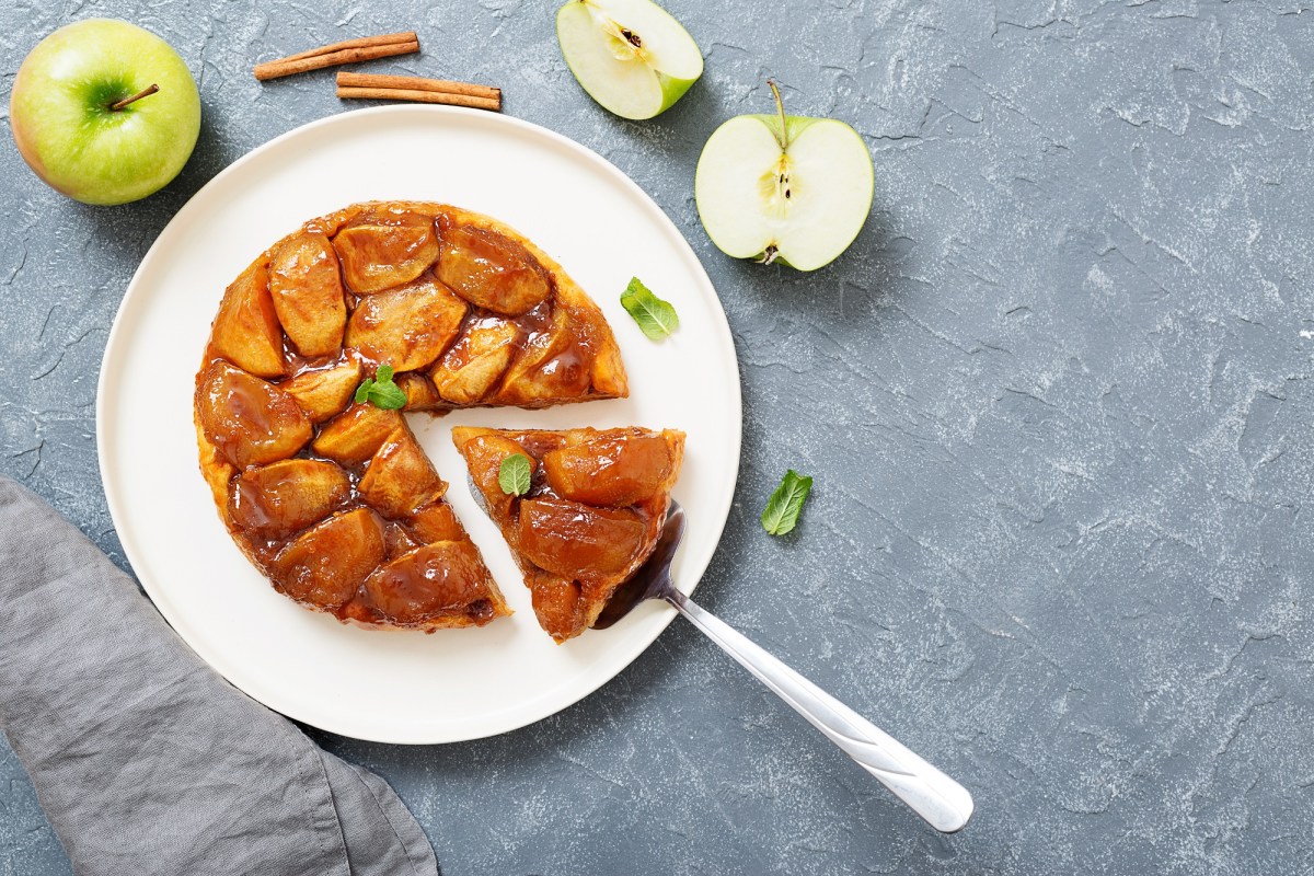 Tarte Tatin auf einem weiÃŸen Teller. Es wurde bereits ein StÃ¼ck abgeschnitten. Daneben liegen angeschnittene grÃ¼ne Ã„pfel und ein paar Zimtstangen.