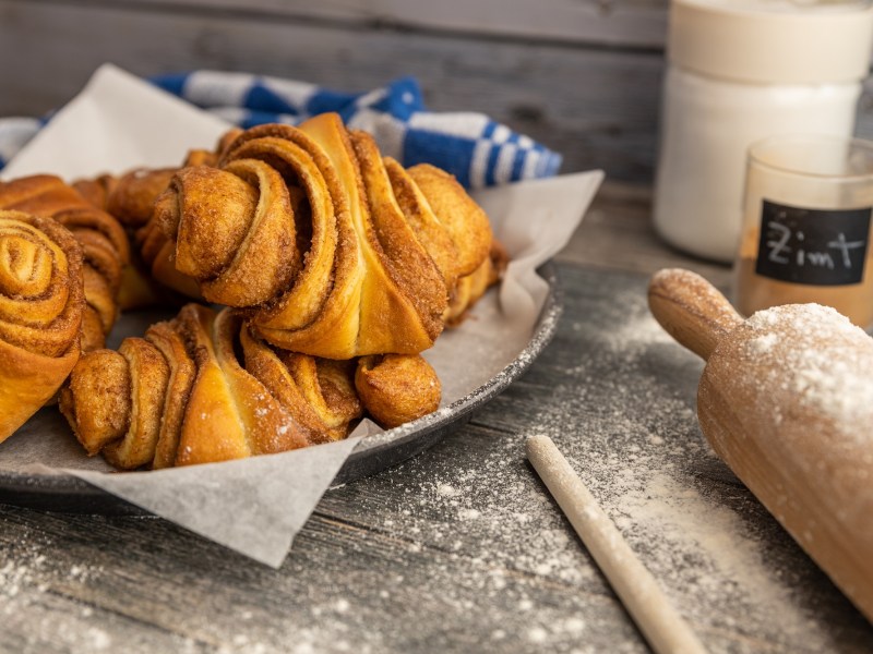 Ein paar vegane FranzbrÃ¶tchen auf einem tiefen Teller, daneben ein Nudelholz, ein Glas mit Zimt und eins mit Zucker.