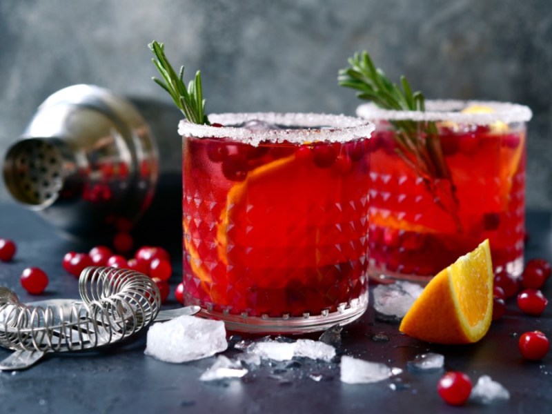 2 GlÃ¤ser Winter-Margarita mit Rosmarinzwigen. Im Hintergrund ein Cocktailshaker, EiswÃ¼rfel, Cranberrys, Orangen und Rosmarinzweige.